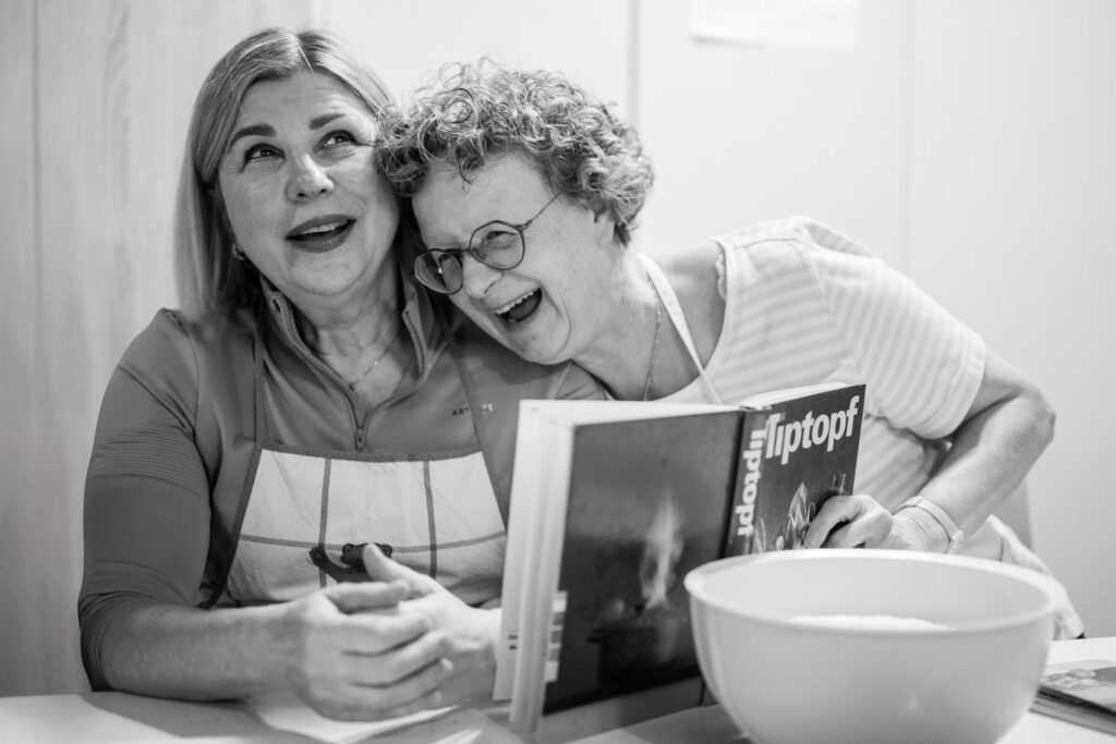 Zwei Frauen sitzen lachend nebeneinander in einer Küche und schauen gemeinsam in das Kochbuch „Tiptopf“, einen klassischen Schweizer Küchenratgeber.