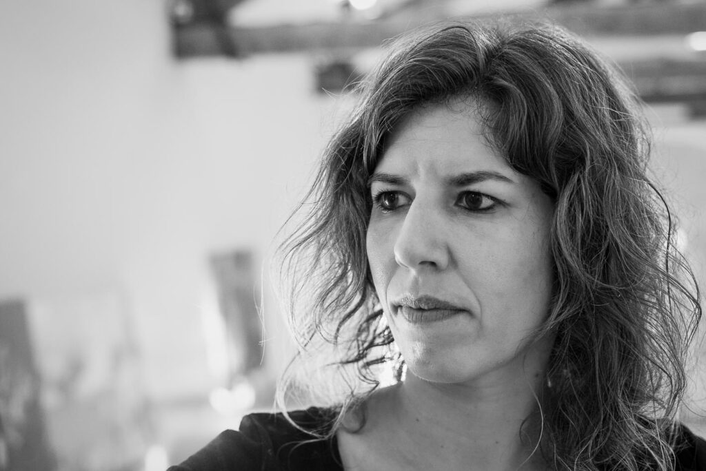 Schwarz-Weiß-Foto einer Frau mit gewelltem Haar, die mit ausdruckslosem Gesicht nach links blickt, aufgenommen in einem sanft beleuchteten Innenraum.