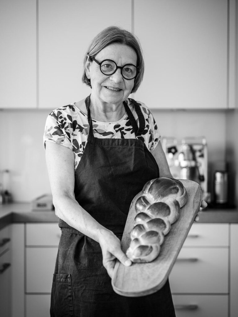 Eine ältere Frau mit Brille und Schürze steht in einer Küche, lächelt und hält ein Holzbrett mit einem Zopf in der Hand.