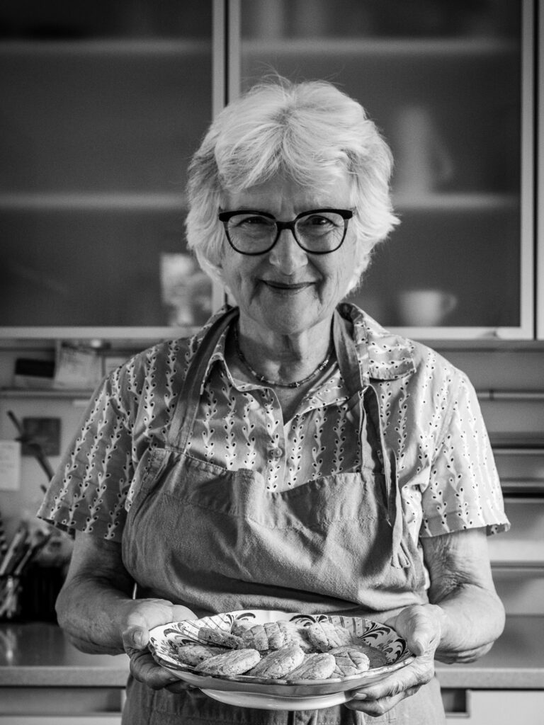 Eine Frau mit weißem Haar, Brille und Schürze zeigt lächelnd eine Platte mit Guetzli.
