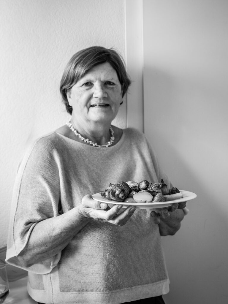 Eine Frau mit einer Halskette und einem hellen Oberteil steht vor einer weißen Wand. Sie hält einen Teller mit einem Gericht aus Gemüse und Fleisch vor sich.